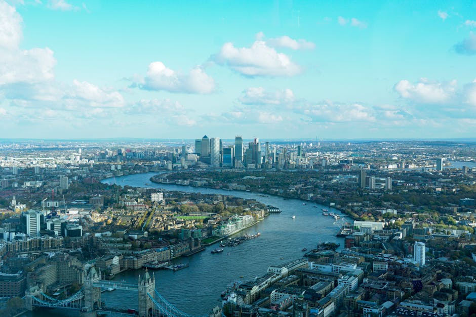 A high-angle view of the London skyline featuring the River Thames winding through the city, with various buildings, bridges, and boats visible along the waterway. The foreground shows dense urban lan