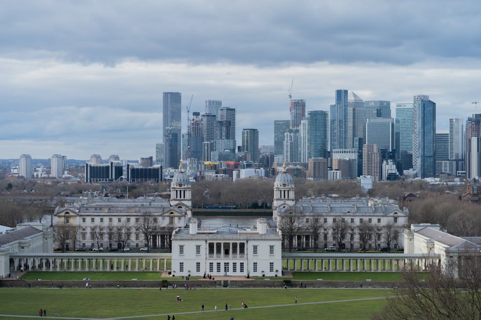 A high-angle view of the London skyline featuring the River Thames winding through the city, with various buildings, bridges, and boats visible along the waterway. The foreground shows dense urban lan