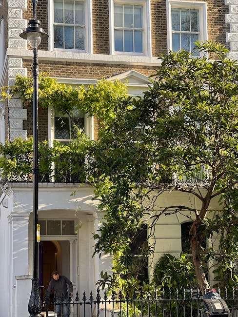 A residential garden scene in Cubitt Town showing a lush, leafy tree with dark green foliage partially obscuring the white-painted façade of a multi-storey brick building. The garden area features a n